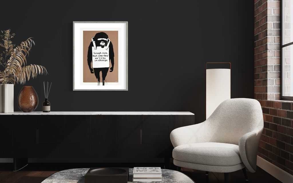 A framed image of a chimpanzee wearing a sandwich board hangs in a dark room beside a pale armchair.