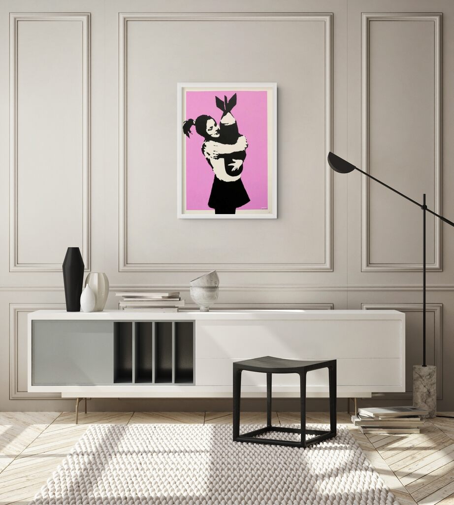 A framed pink print of a girl embracing a bomb hangs above a white sideboard in a pale panelled room.
