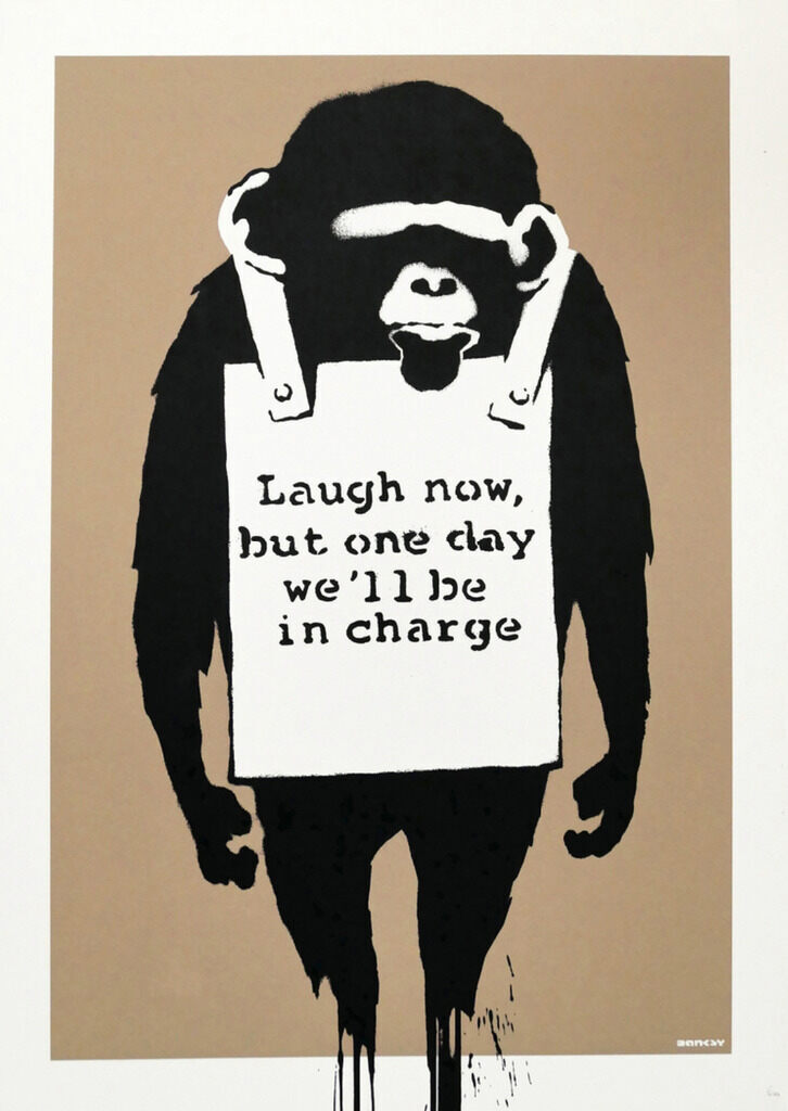 Uma figura de macaco preto está de pé com ambos os braços pendurados ao lado do corpo, vestindo uma placa de anúncio (sandwich board) branca com as palavras "Laugh now, but one day we'll be in charge" sobre um fundo marrom.