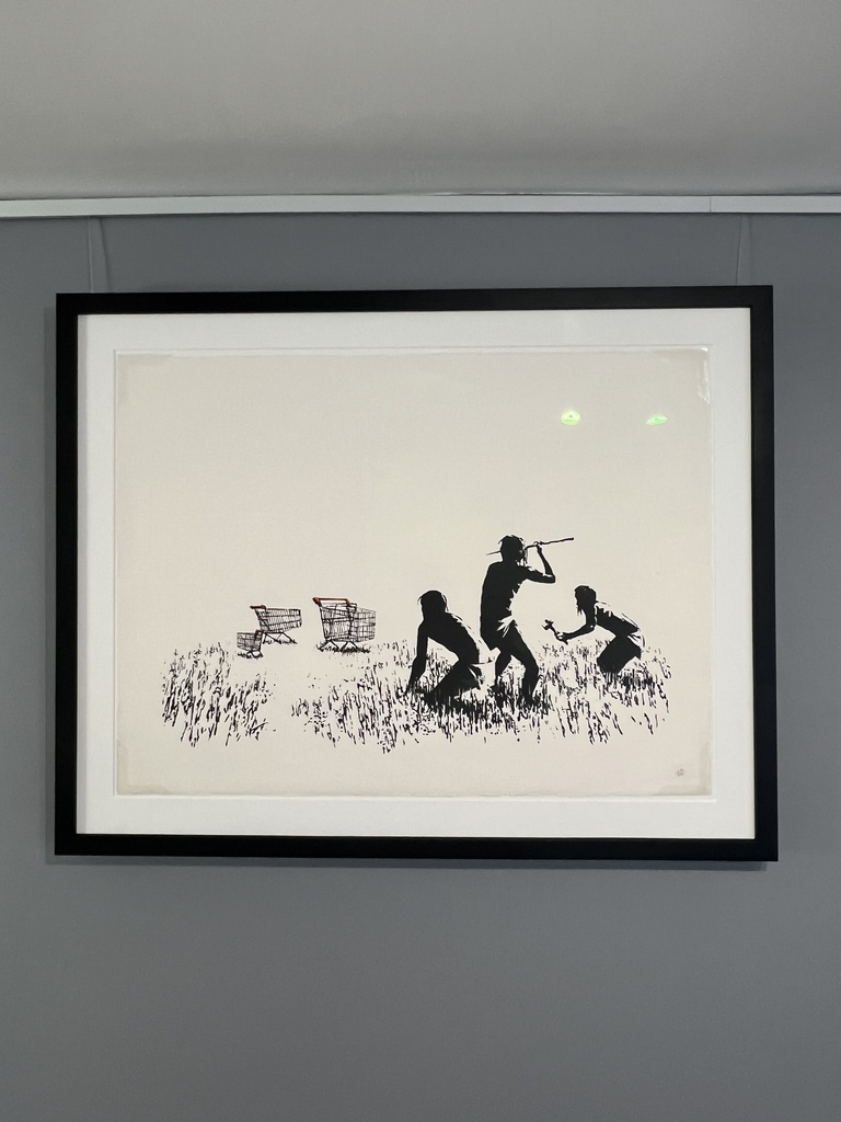 Three dark figures stand and crouch in grass opposite three shopping trolleys; the central figure holds a spear above his shoulder.