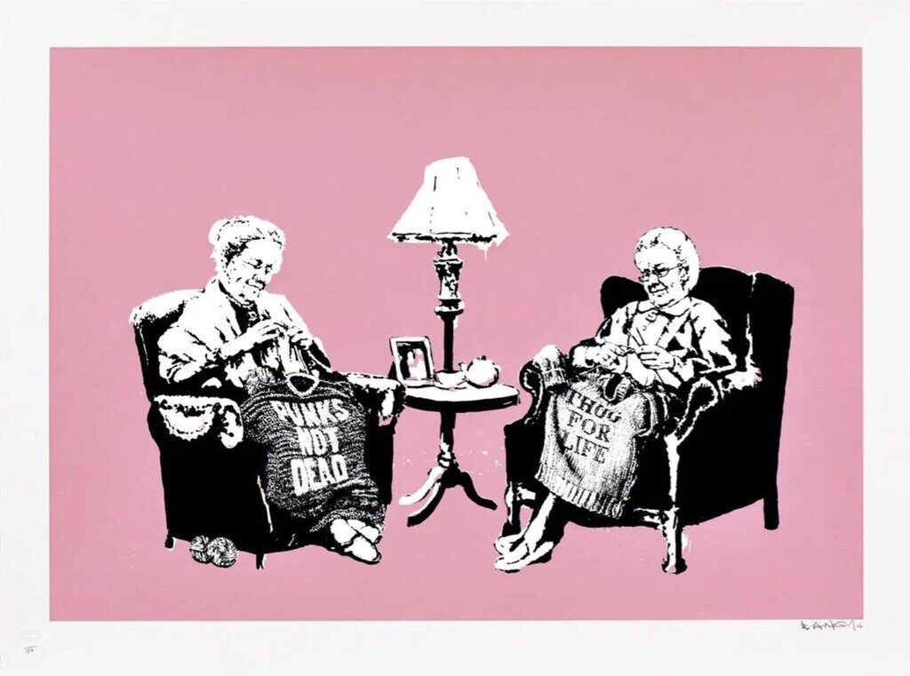 Two elderly women sit in armchairs knitting jumpers bearing PUNKS NOT DEAD and THUG FOR LIFE, with a small table and lamp between them, against pink.