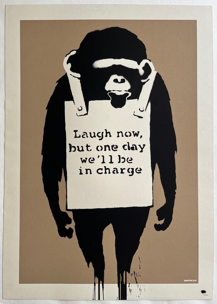 A black ape figure stands with both arms hanging at its sides, wearing a white sandwich board bearing the words "Laugh now, but one day we'll be in charge" against a brown background.