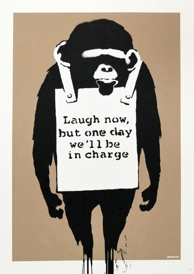 Uma figura de macaco preto está de pé com ambos os braços pendurados ao lado do corpo, vestindo uma placa de anúncio (sandwich board) branca com as palavras "Laugh now, but one day we'll be in charge" sobre um fundo marrom.