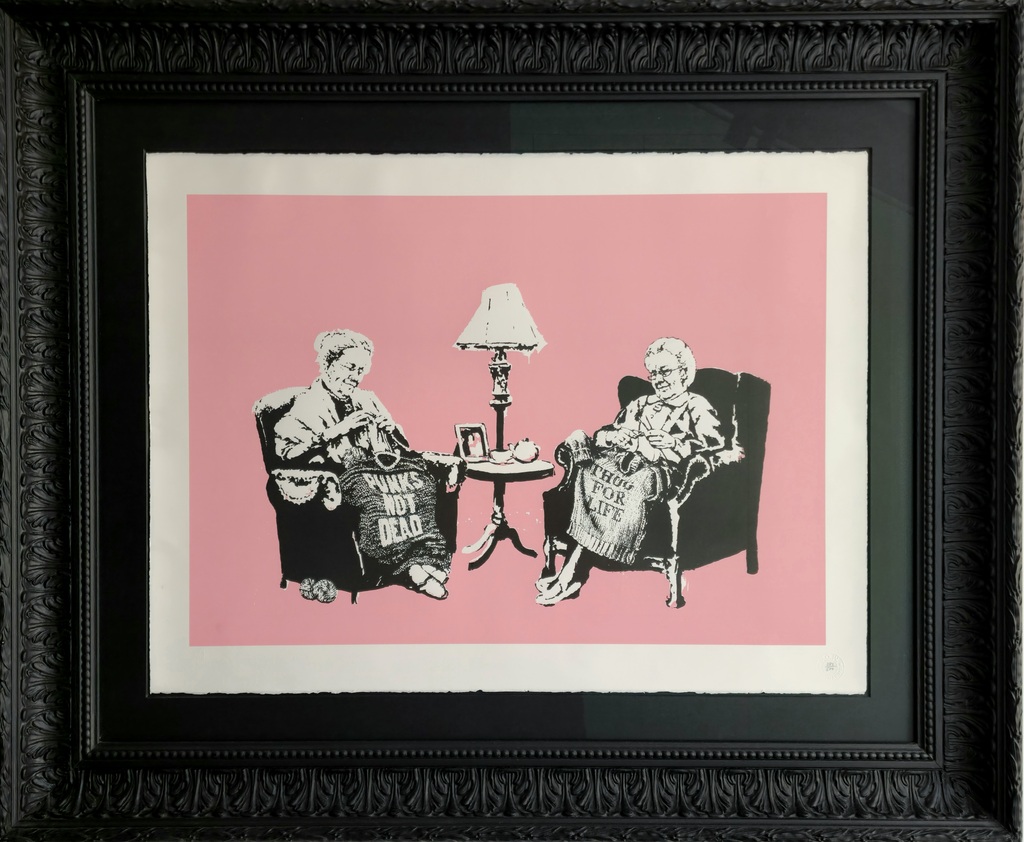 Två äldre kvinnor sitter i fåtöljer och stickar tröjor med texten PUNKS NOT DEAD och THUG FOR LIFE, med ett litet bord och en lampa mellan sig, mot en rosa bakgrund.