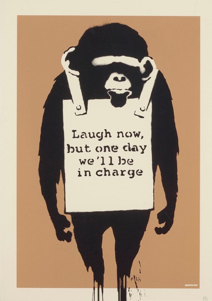 Figura de un simio negro de pie con ambos brazos colgando a los lados, vistiendo un cartel publicitario de sándwich con las palabras "Laugh now, but one day we'll be in charge" sobre un fondo marrón.